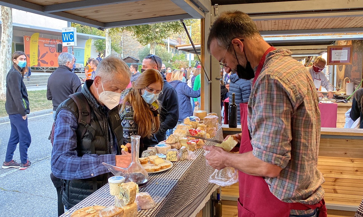 Pla mitjà d'un formatger tallant formatge a la seva parada i dos visitants de la Fira de Sant Ermengol de la Seu d'Urgell a punt per degustar-lo. Imatge publicada el 17 d'octubre de 2021. (Horitzontal)